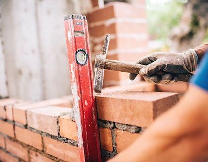 A gloved hand uses a brick hammer to adjust a brick next to a vertical red spirit level on a construction site.