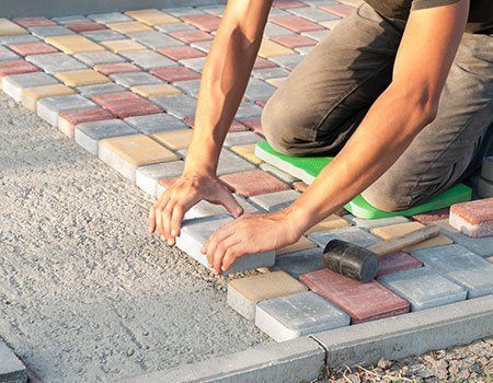 A person kneeling on a green pad while laying multicolored rectangular pavers on a prepared gravel base.