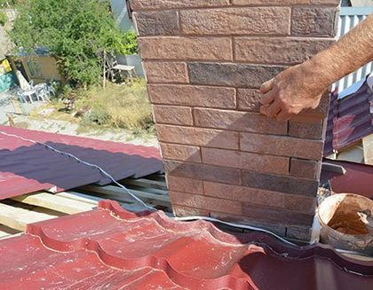 A person’s hand applies mortar to a brick chimney stack on a red metal roof.