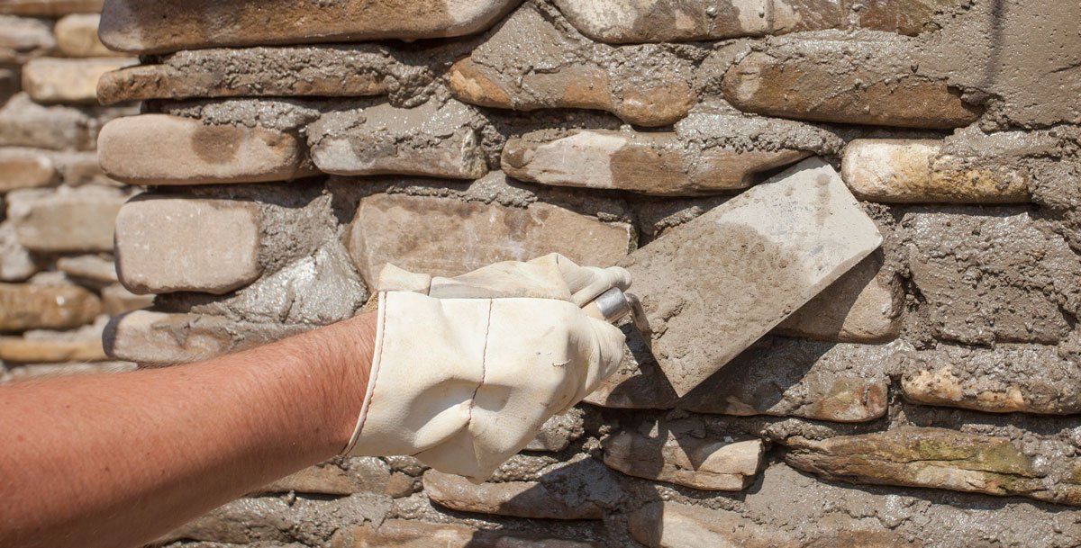A gloved hand presses a flat stone into a layer of wet mortar on a rustic stone wall.