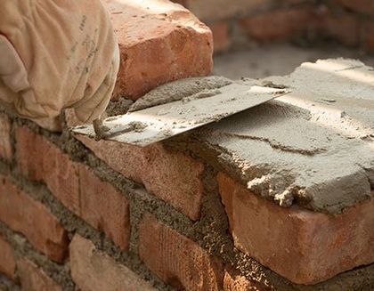 A gloved hand uses a metal trowel to spread gray mortar over a layer of red clay bricks during construction.