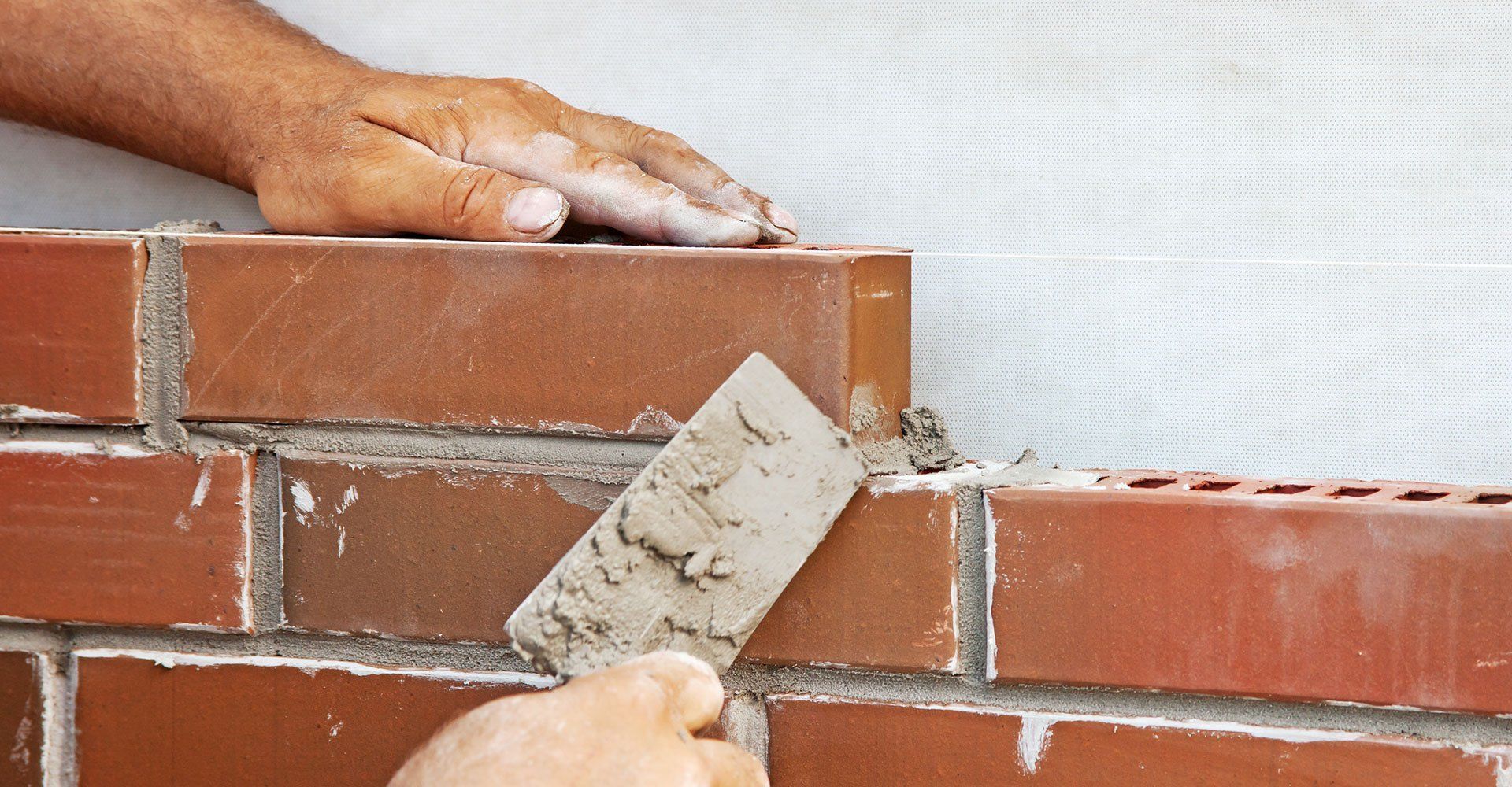 A mason uses a metal trowel to apply mortar between red bricks on a construction site.