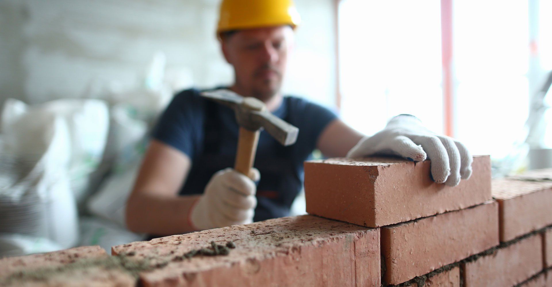 A construction worker in a yellow hard hat and gloves uses a hammer to lay a red brick on a wall.