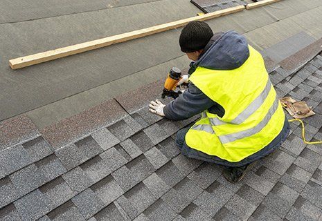 A roofer in a yellow safety vest uses a nail gun to install grey asphalt shingles on a sloped roof.