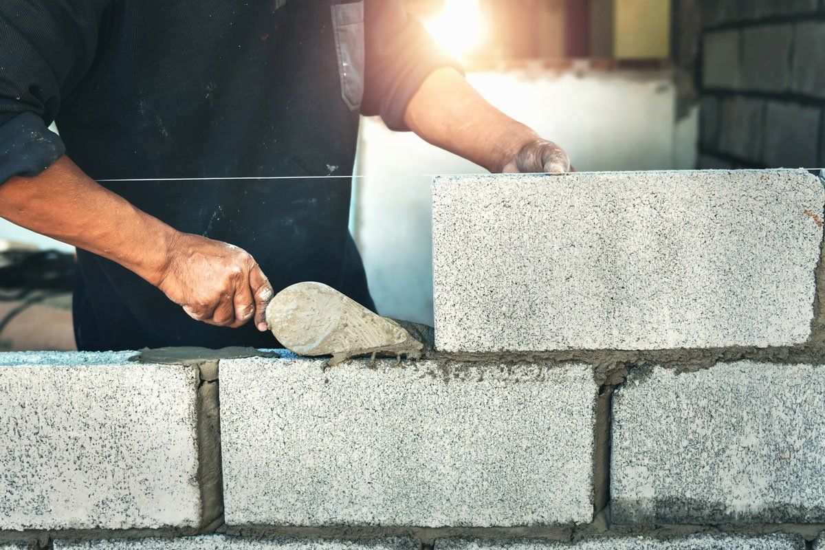 A construction worker uses a metal trowel to spread mortar onto grey concrete blocks while building a wall.