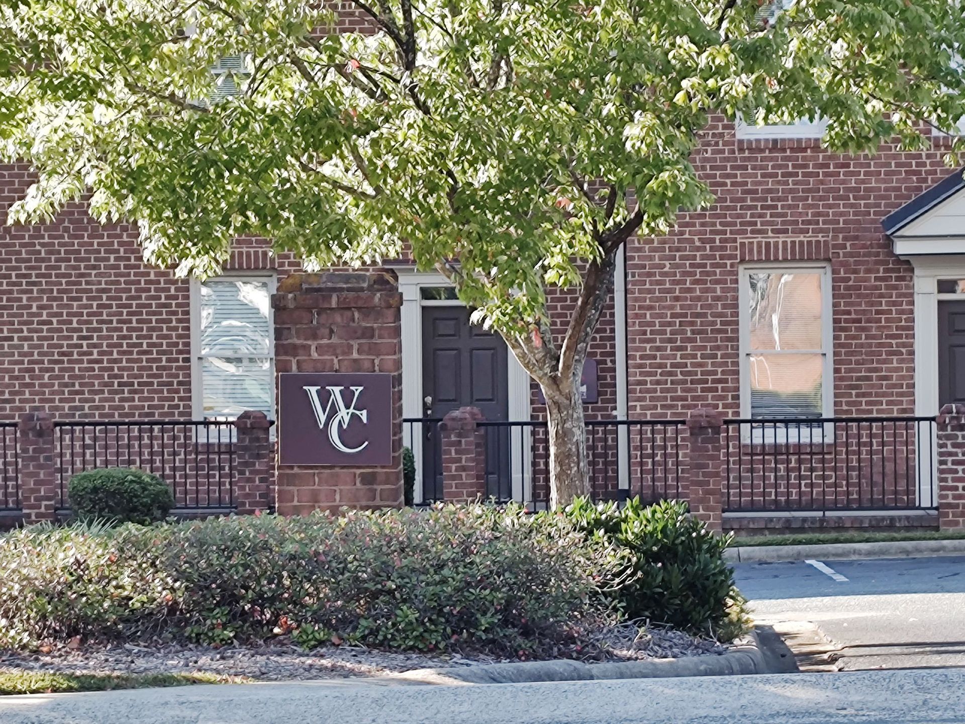 A brick office building with a "WC" sign in front, partially obscured by a tree, with a paved parking area in the foreground.