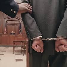 A person in a suit is handcuffed behind their back inside a courtroom, with another person's hand resting on their arm.