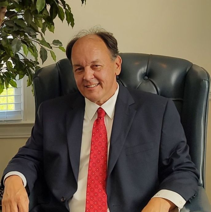 A person in a dark blue suit and red tie sits in a tufted leather chair next to a green plant.
