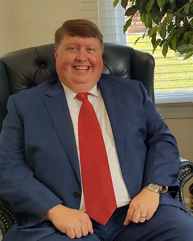 A person wearing a blue suit and red tie sits in a black leather armchair, smiling at the camera in a brightly lit room.