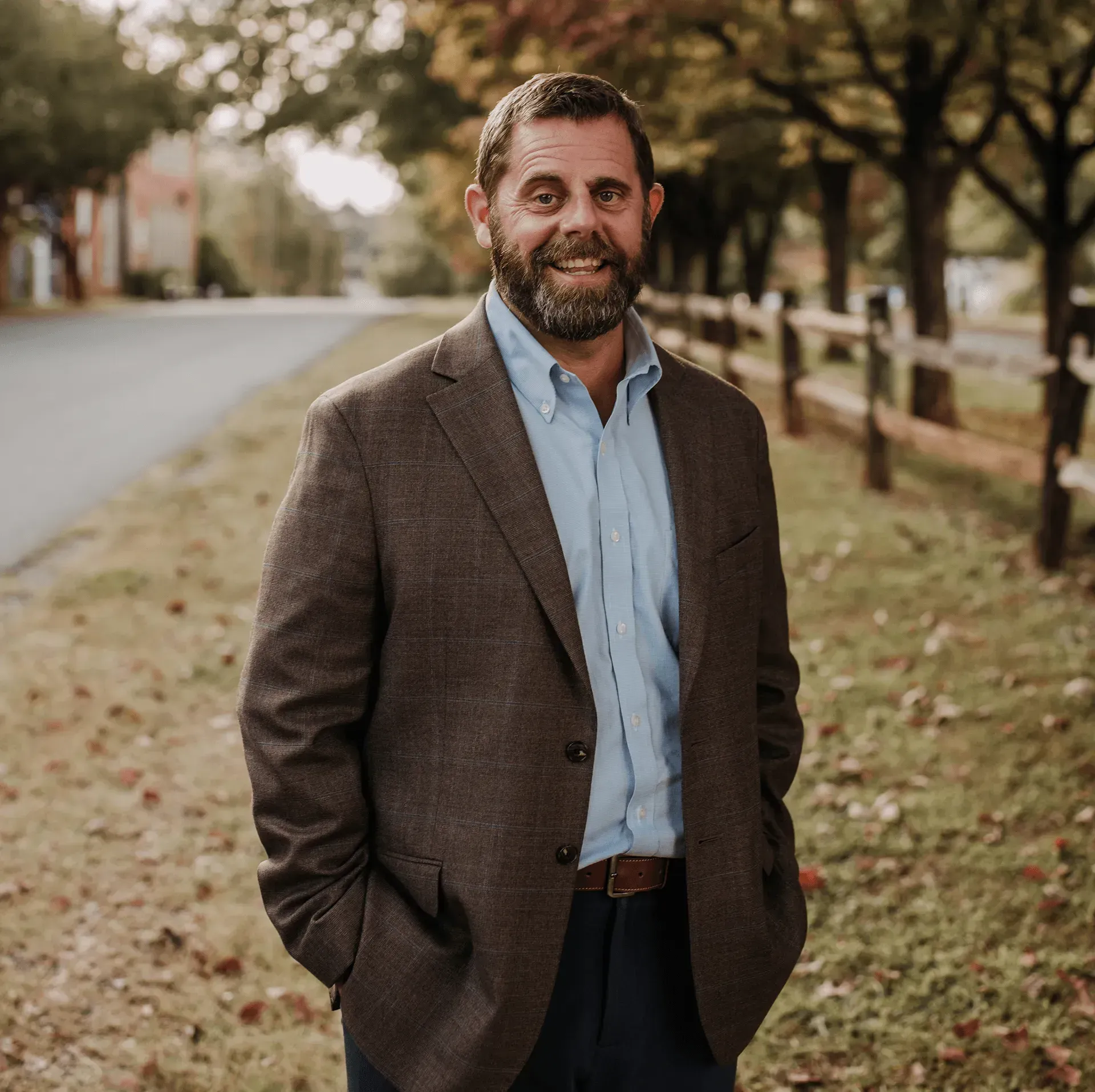 A person with a beard, wearing a brown blazer and blue shirt, stands smiling on a path near a wooden fence.