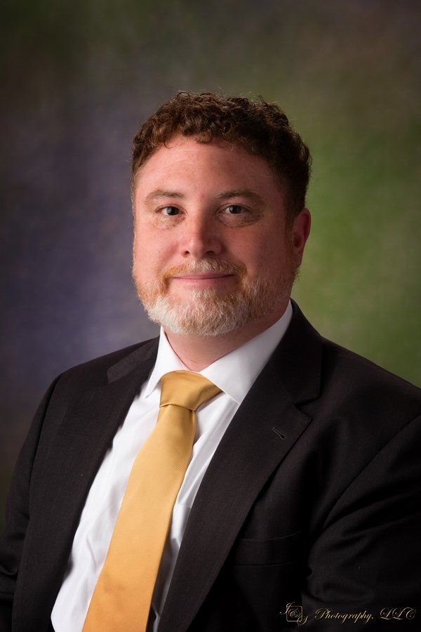 A person with short wavy hair, a beard, and a yellow tie, wearing a dark suit jacket against a mottled green backdrop.
