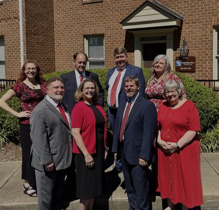 Eight people in business attire pose for a photo in front of a brick building entrance surrounded by green shrubs.