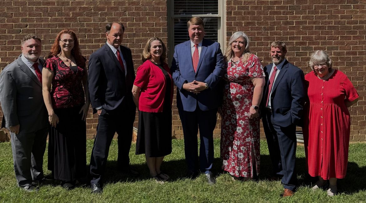 A group of eight people in formal attire pose for a photo in front of a brick building.