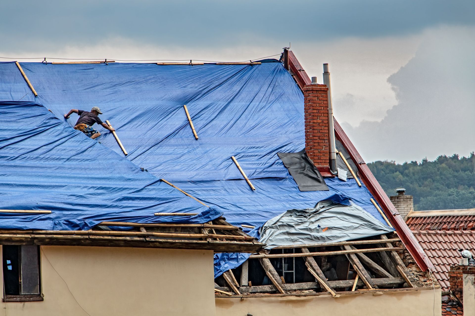 A person secures a blue tarp over a damaged, partially open roof with exposed wooden rafters and a brick chimney.