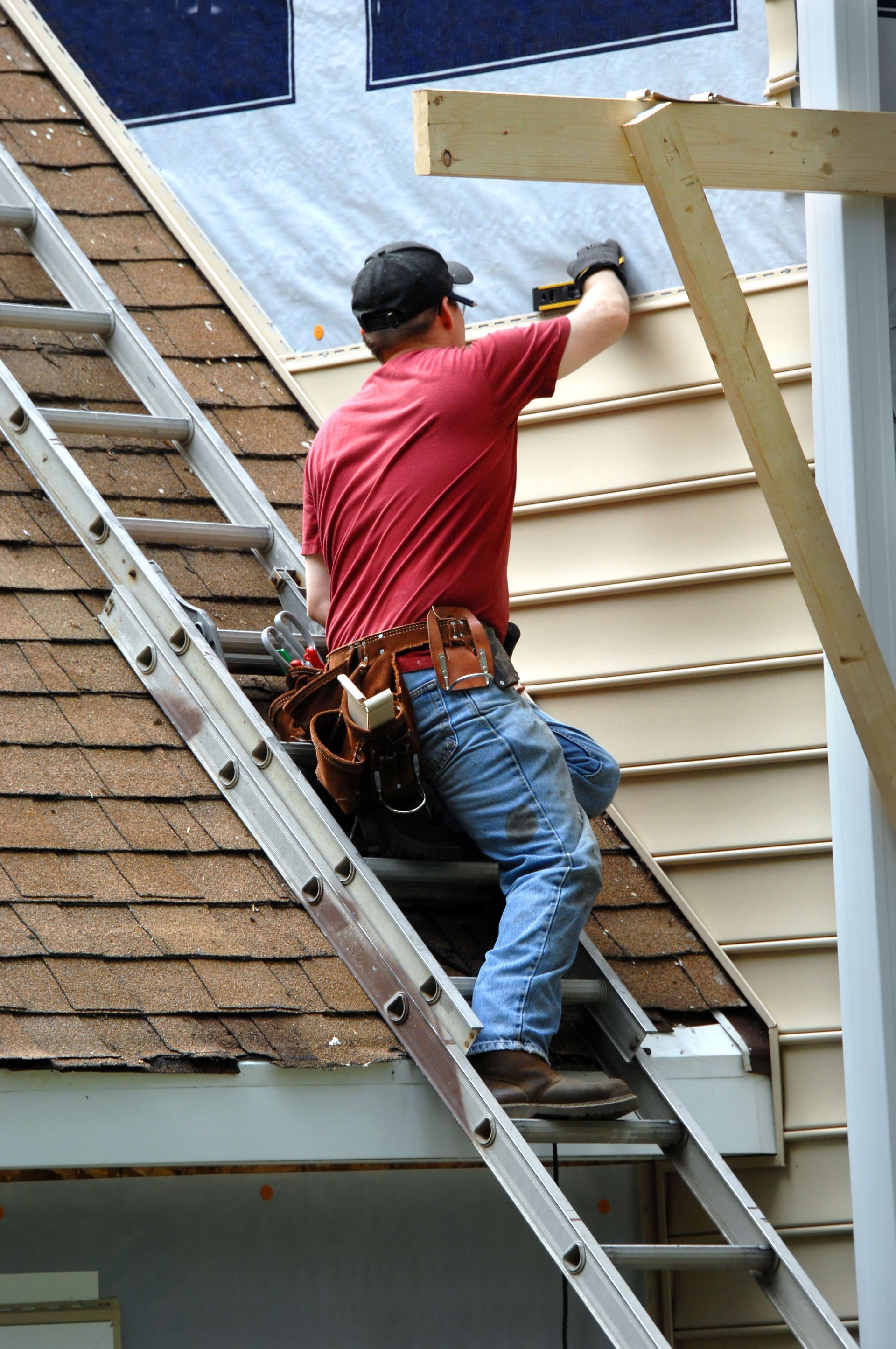 A person wearing a red shirt and tool belt stands on a ladder, installing horizontal siding on a house exterior.