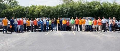 A large group of people in colorful shirts stands in a parking lot, smiling and posing in front of a row of vehicles.