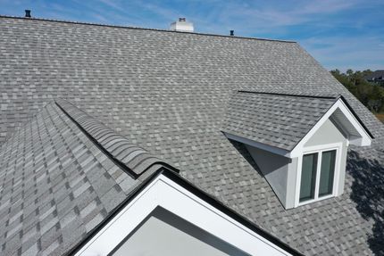 Aerial view of a residential shingled roof with a dormer and two gable sections against a blue sky.