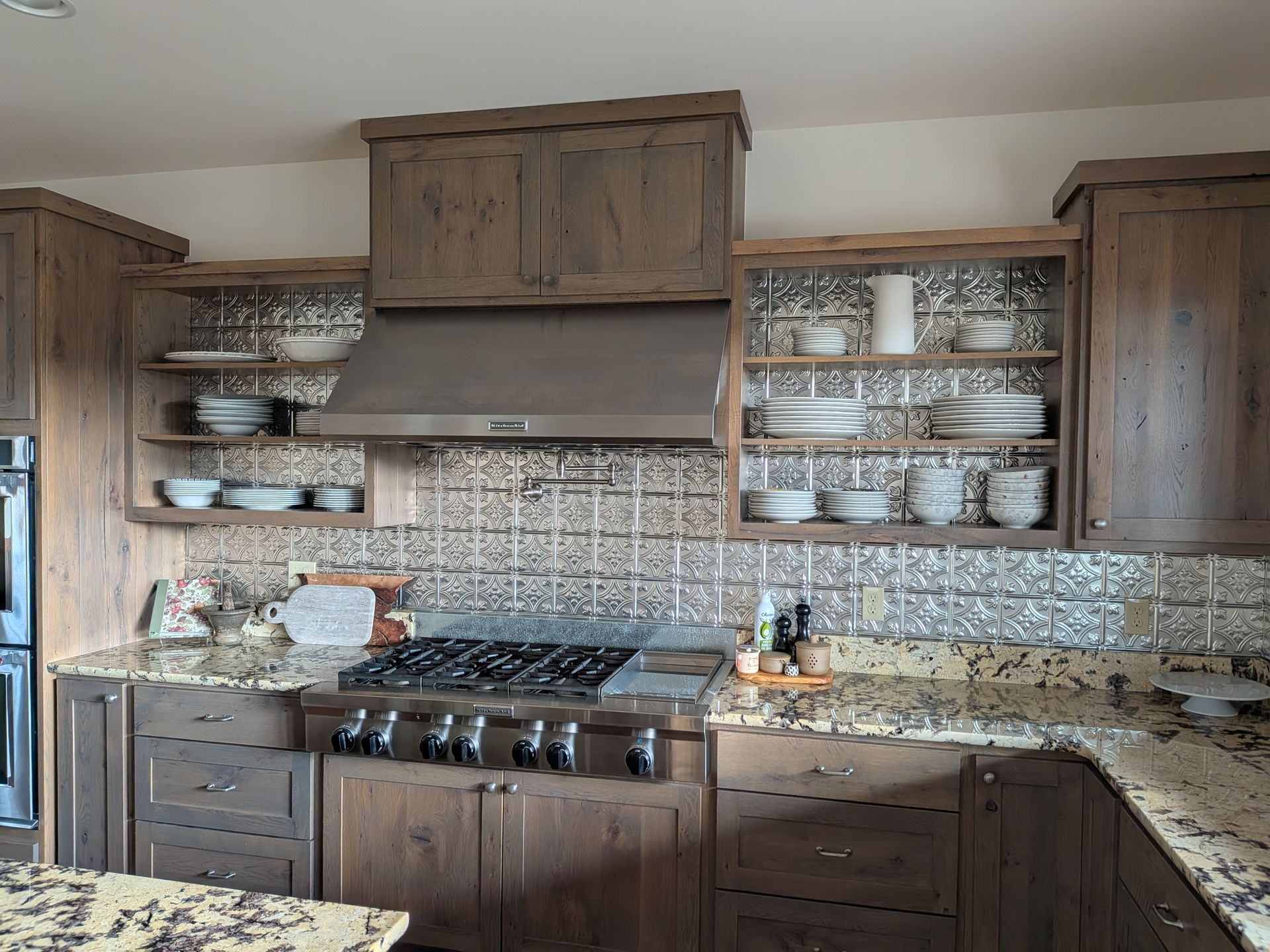 Rustic kitchen with wooden cabinets, tiled backsplash, open shelves, and a large stainless range hood.