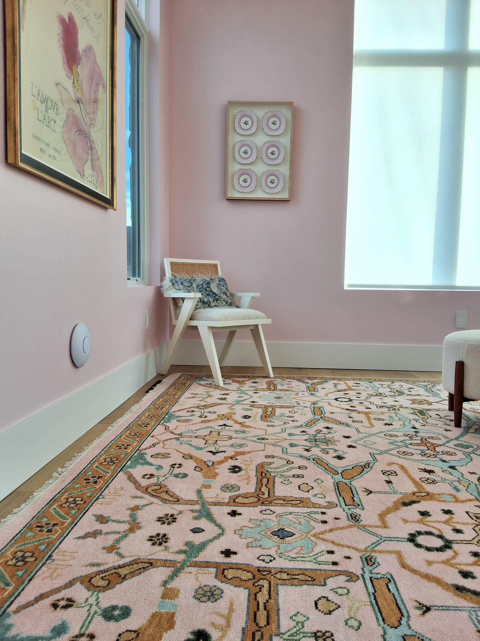 Empty pastel room with patterned rug, chair, wall art, and bright window.