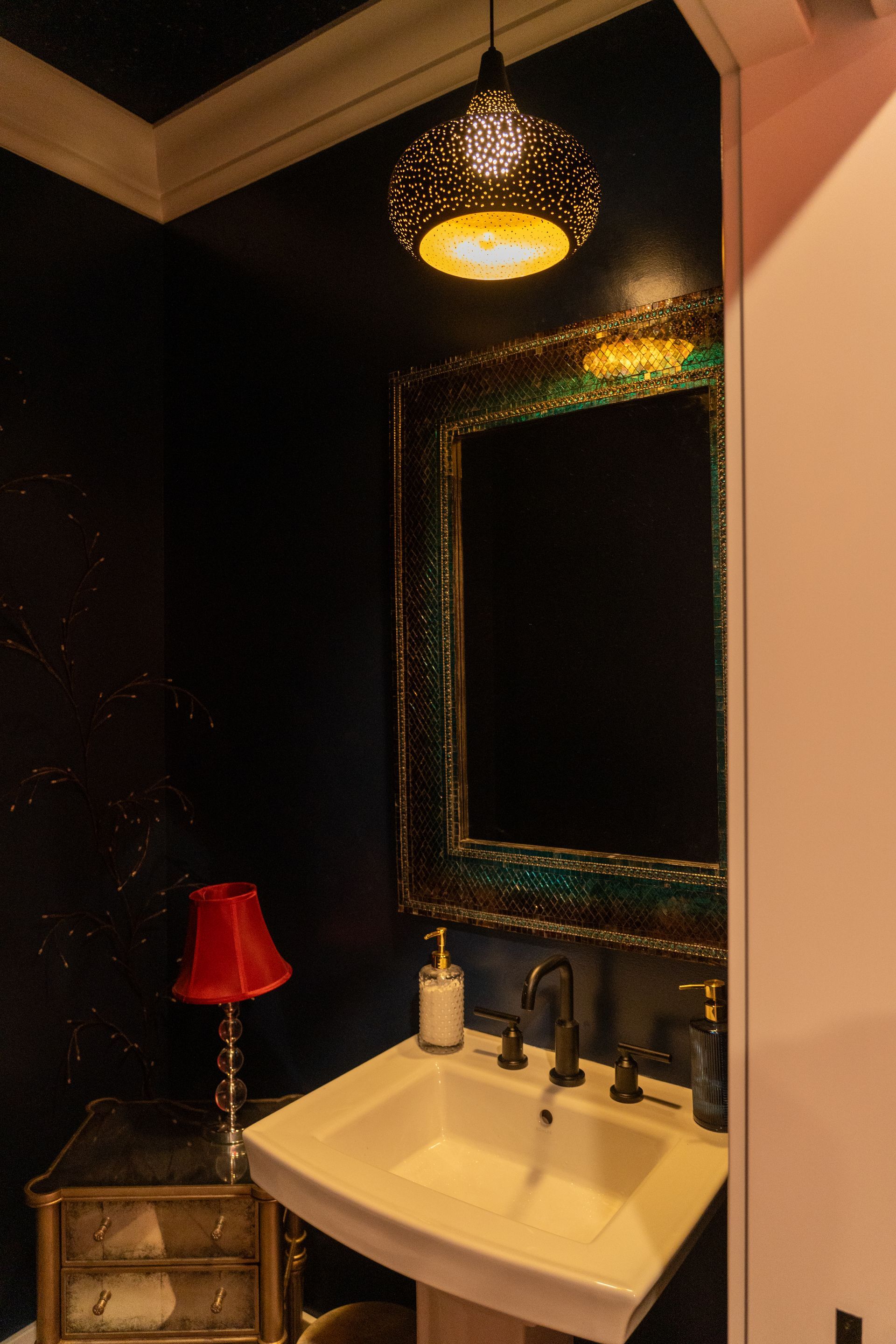 Small bathroom vanity with dark walls, ornate mirror, black faucet, and red lamp on a side table.