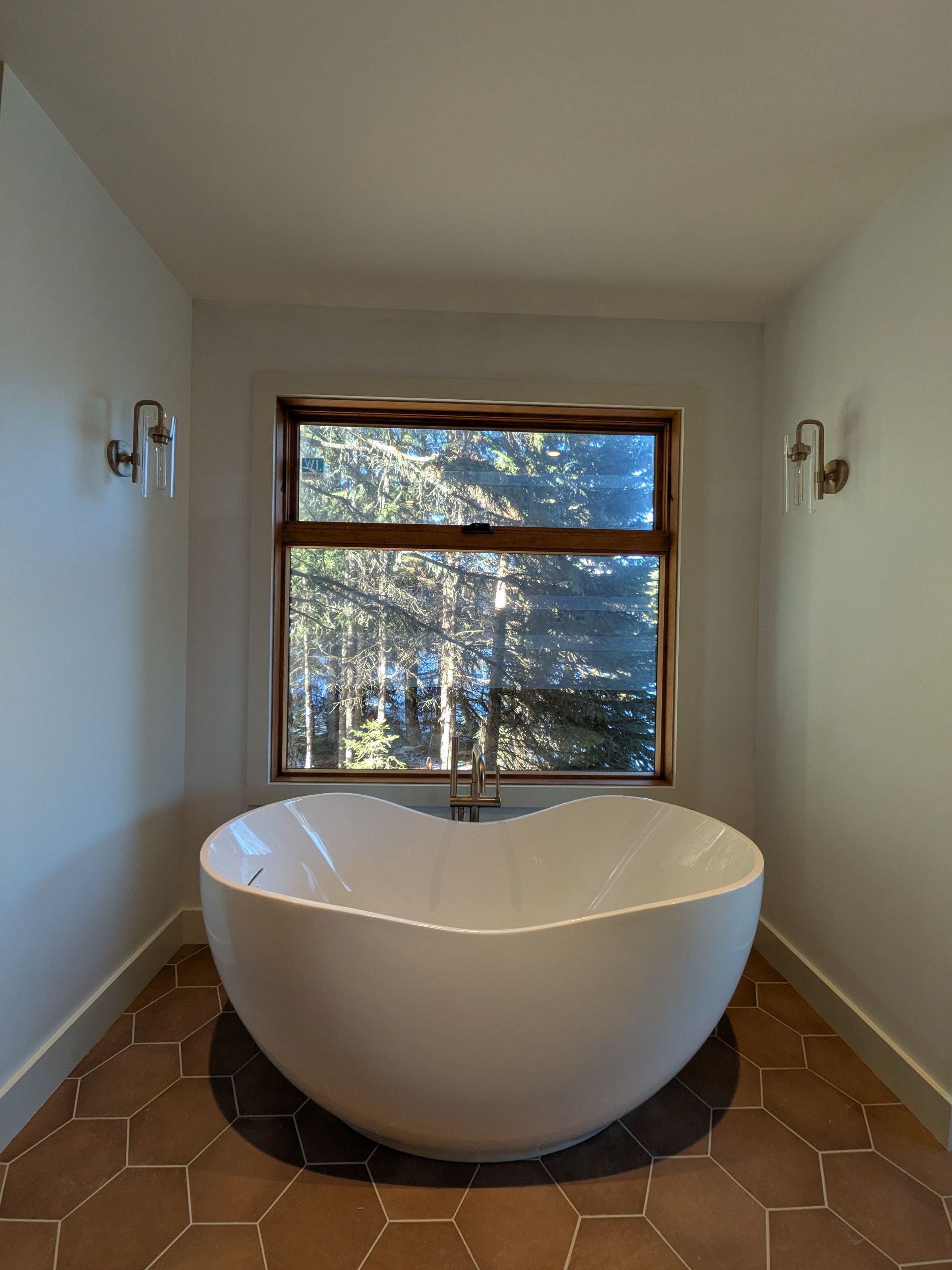 Modern freestanding white bathtub in a narrow room with a window and terracotta tile floor
