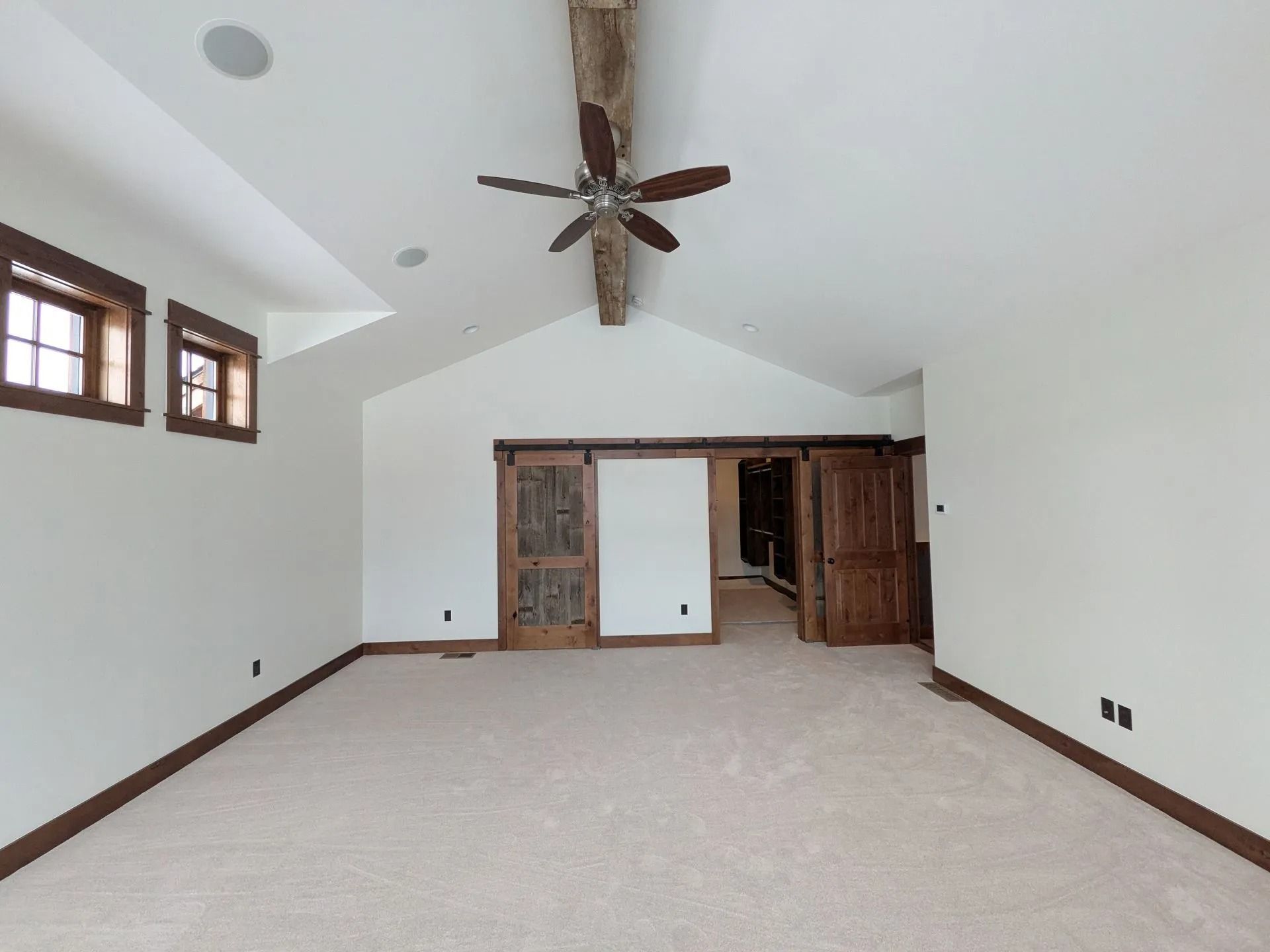 A bright, empty room with white walls, carpeted floors, a vaulted ceiling with a rustic wooden beam, and sliding doors.