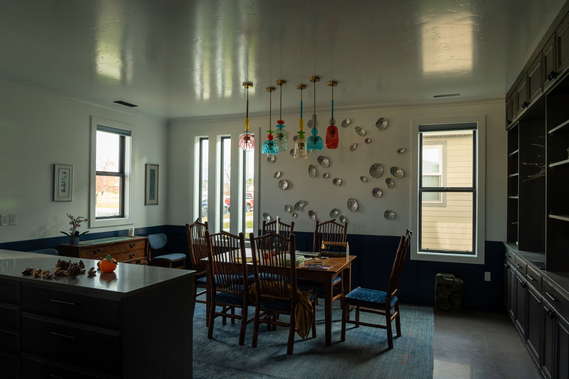 Dining room with a wooden table, chairs, hanging colorful lights, and large windows in a modern home