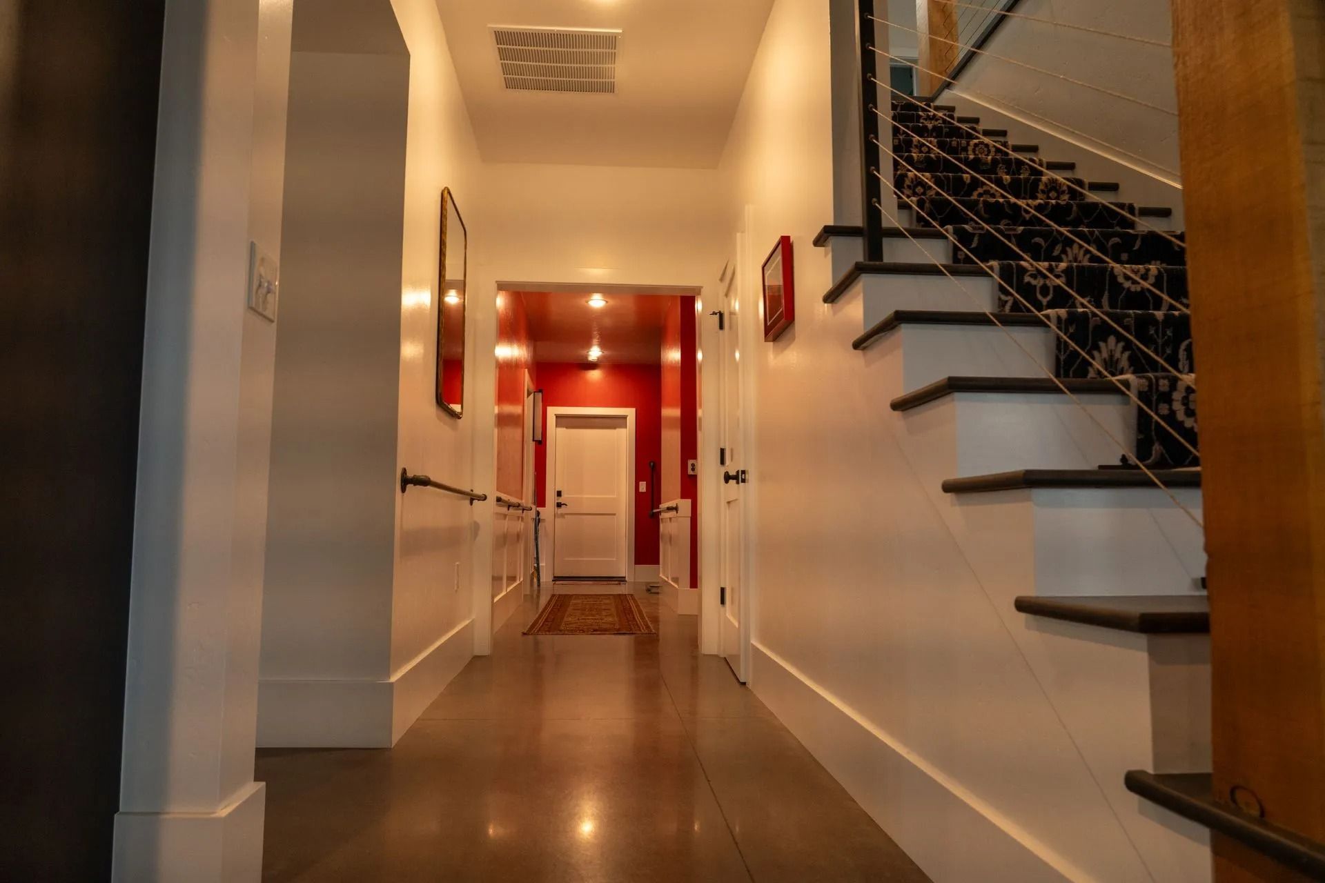 A hallway with white walls and polished floors leads to a bright red room with a white door, alongside a wooden staircase.
