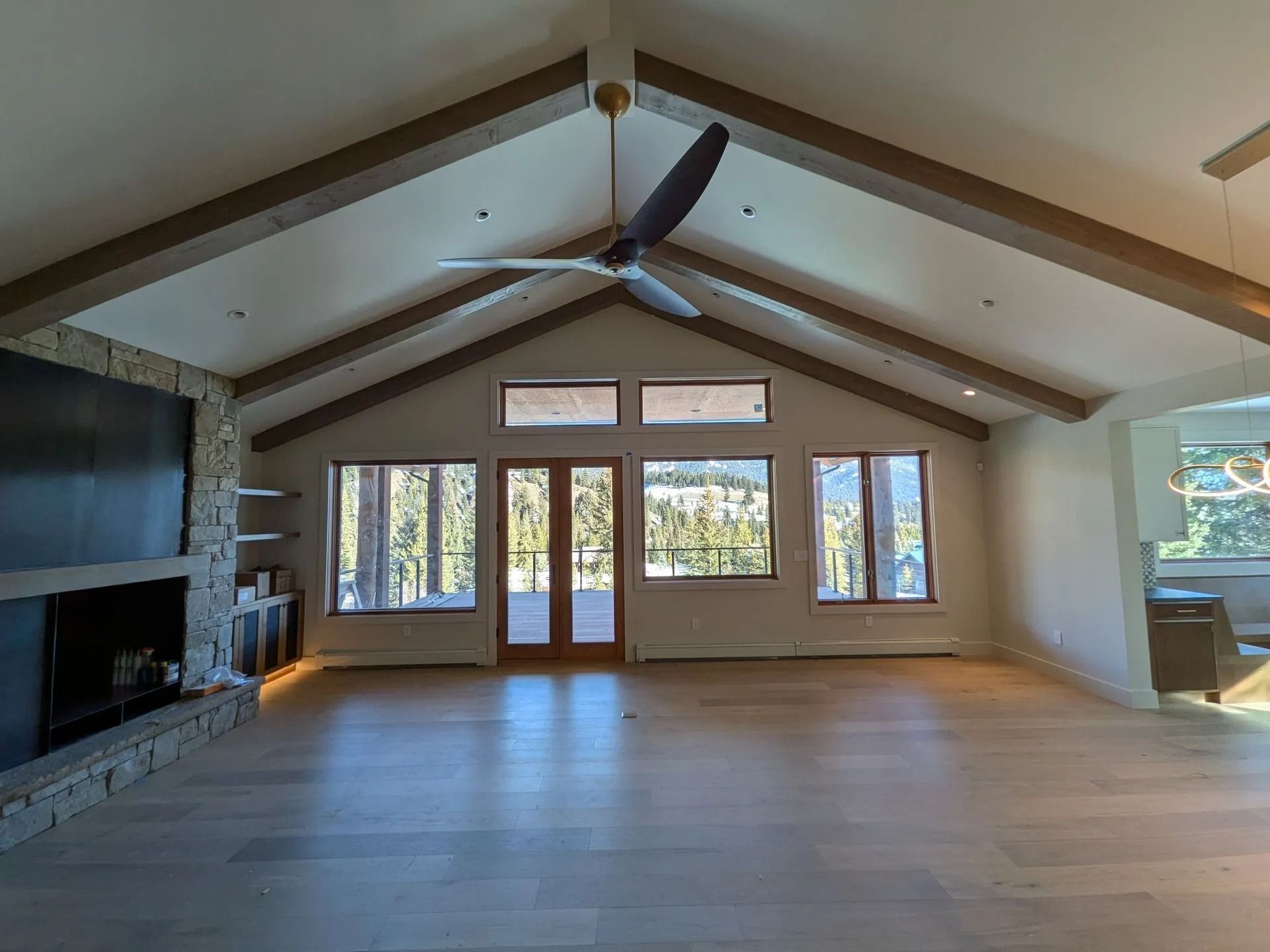 A living room with light wood floors, a vaulted ceiling with wooden beams, a stone fireplace, and large windows.