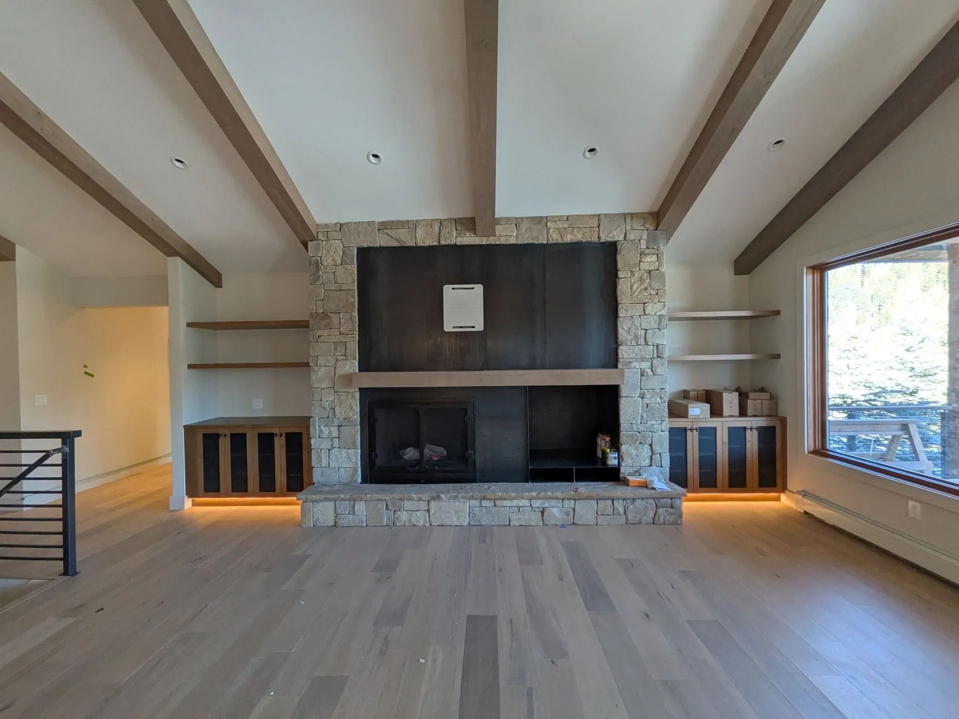 A modern living room with a large stone fireplace, exposed wooden ceiling beams, light wood floors, and built-in shelves.