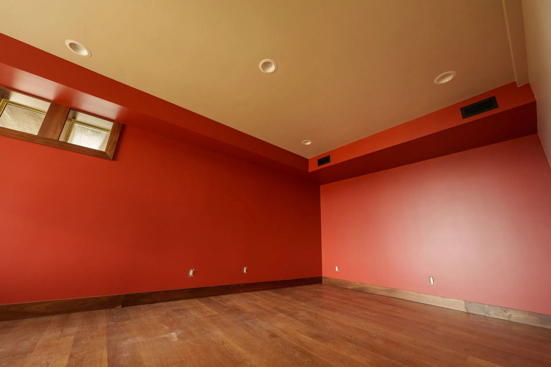 An empty room with bold red walls, brown wood flooring, recessed ceiling lights, and small windows.