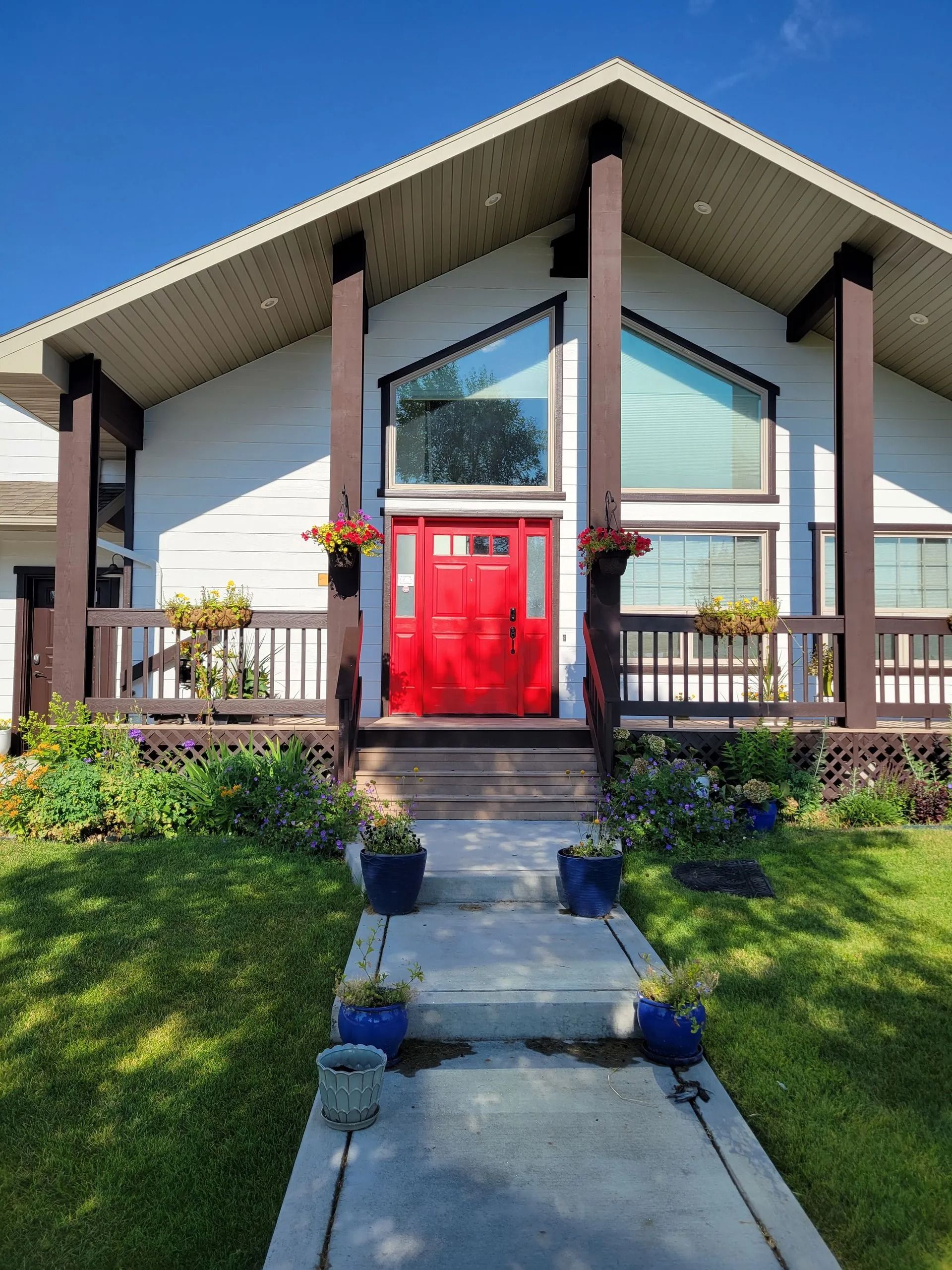 A bright red front door stands beneath a large, triangular window on a house with white siding and dark brown porch beams.