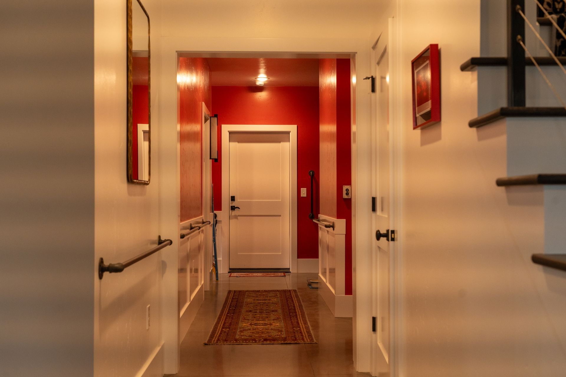 A hallway leads to a red-walled entrance with a white door and a runner rug, flanked by white walls and a staircase.