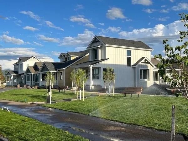 A row of modern, light-colored suburban houses with dark roofs beside a grass lawn and a paved path under a blue sky.