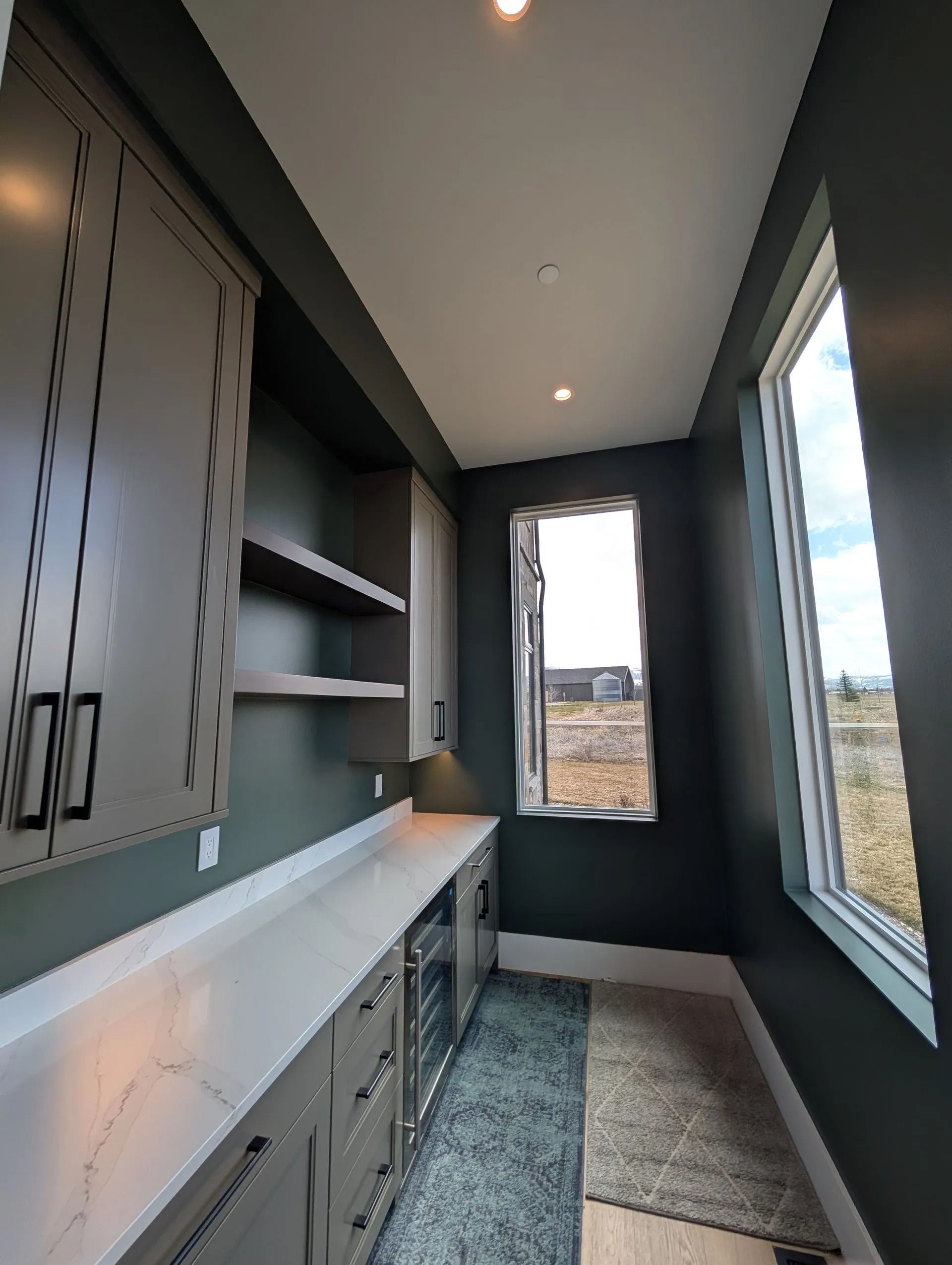 A narrow, dark green galley-style butler’s pantry with white marble counters, shaker cabinets, and two large windows.