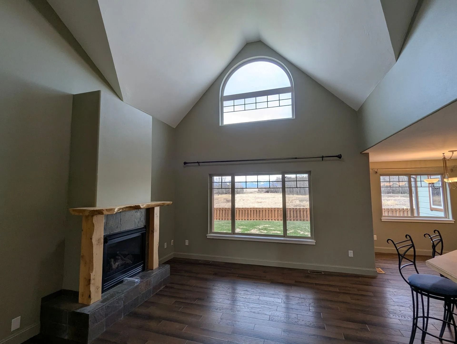 A high-ceilinged room with a rustic fireplace, arched window, and dark wood flooring, overlooking a grassy yard.