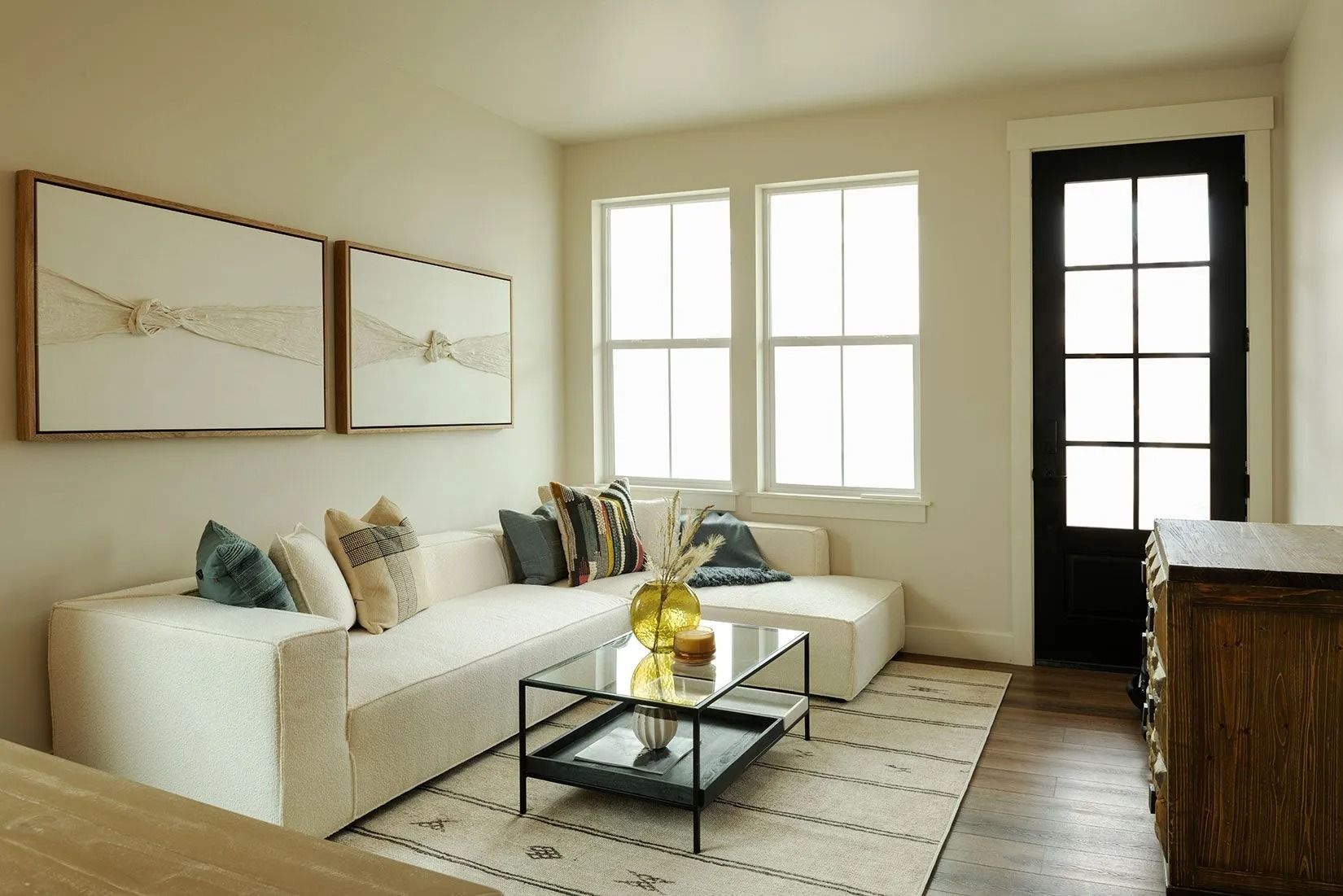A cozy living room with a white sectional sofa, a glass coffee table, patterned area rug, and a black-framed glass door.