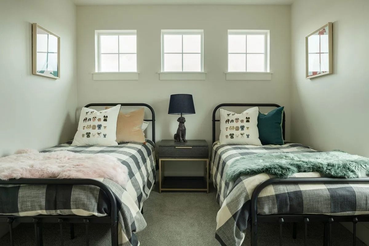 Two twin beds with black metal frames and plaid bedding, separated by a nightstand with a dark lamp, under three windows.