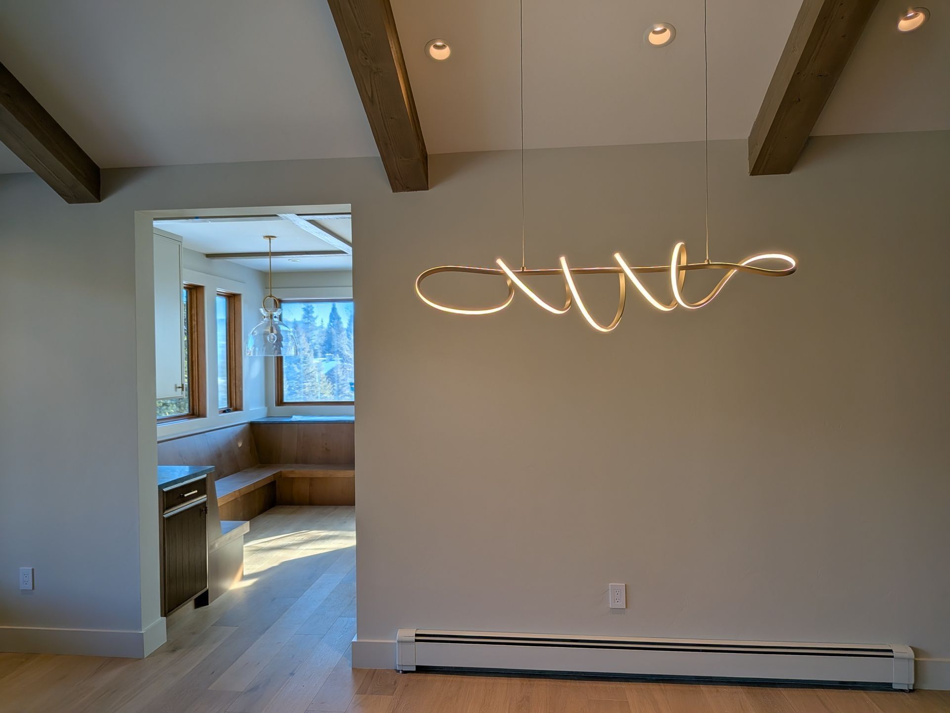 A modern spiral light fixture hangs in a neutral room with wood ceiling beams, looking into a cozy window-seat nook.
