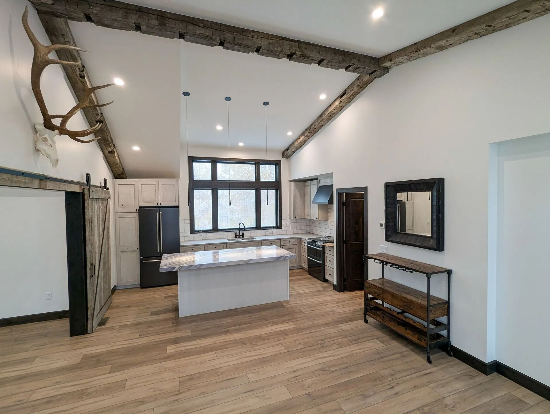Modern kitchen and living area with white walls, rustic wooden ceiling beams, a central island, and wood flooring.
