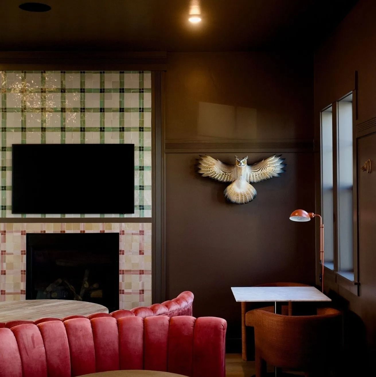 A room with a plaid-tiled fireplace, a wall-mounted owl sculpture, a red velvet curved sofa, and a small cafe table.