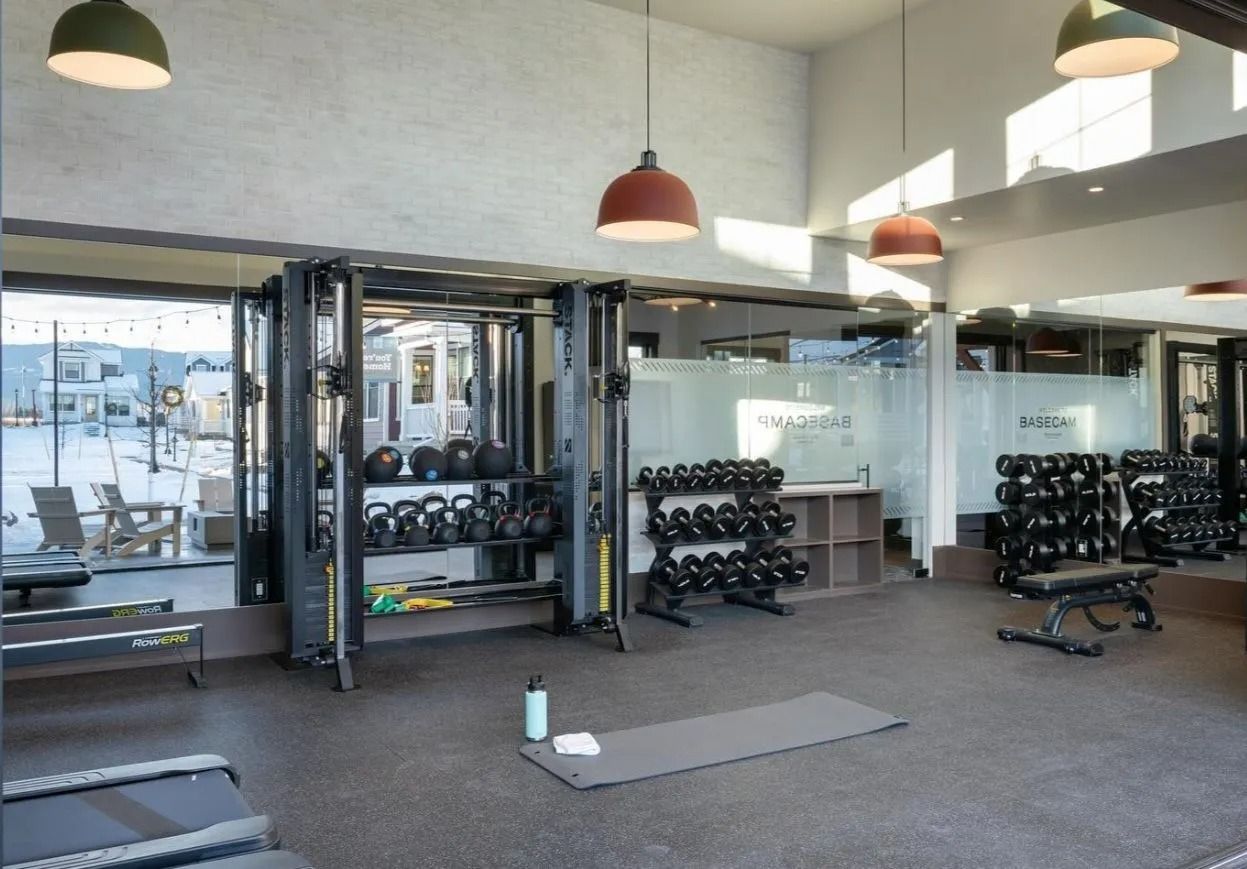 A brightly lit gym with mirrors, a weight rack, dumbbells, a cable machine, and a yoga mat on the floor.