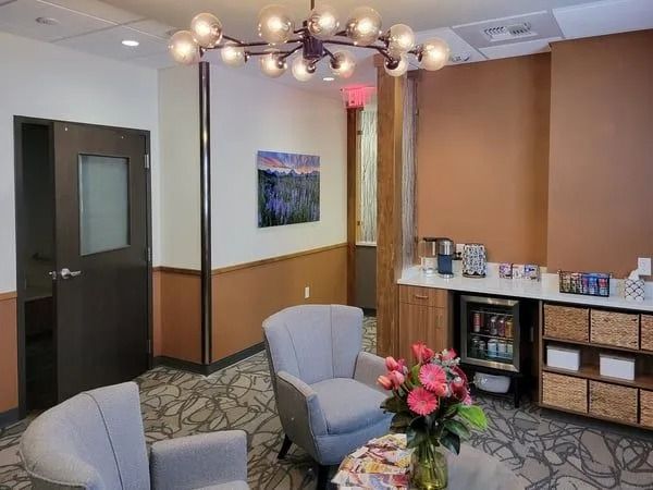 A lounge area with two light gray armchairs, a patterned carpet, a minibar, shelves, and a modern chandelier.