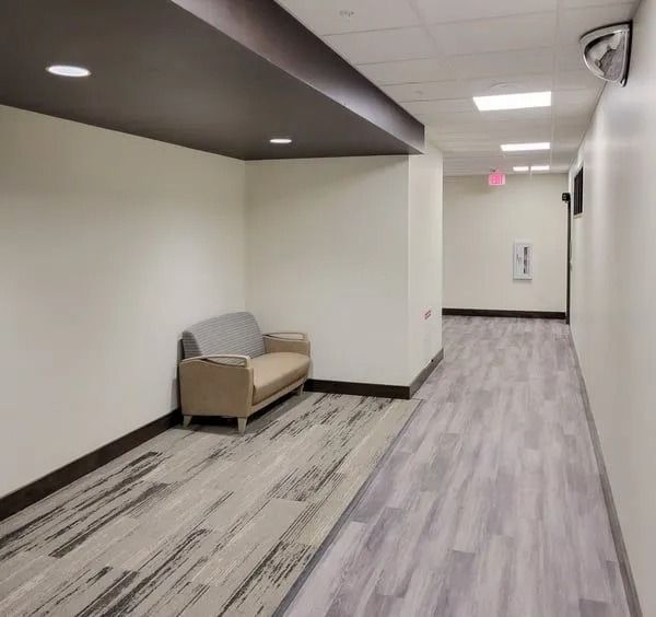 A beige sofa sits in a small, modern office alcove next to a long hallway with light wood-look flooring.