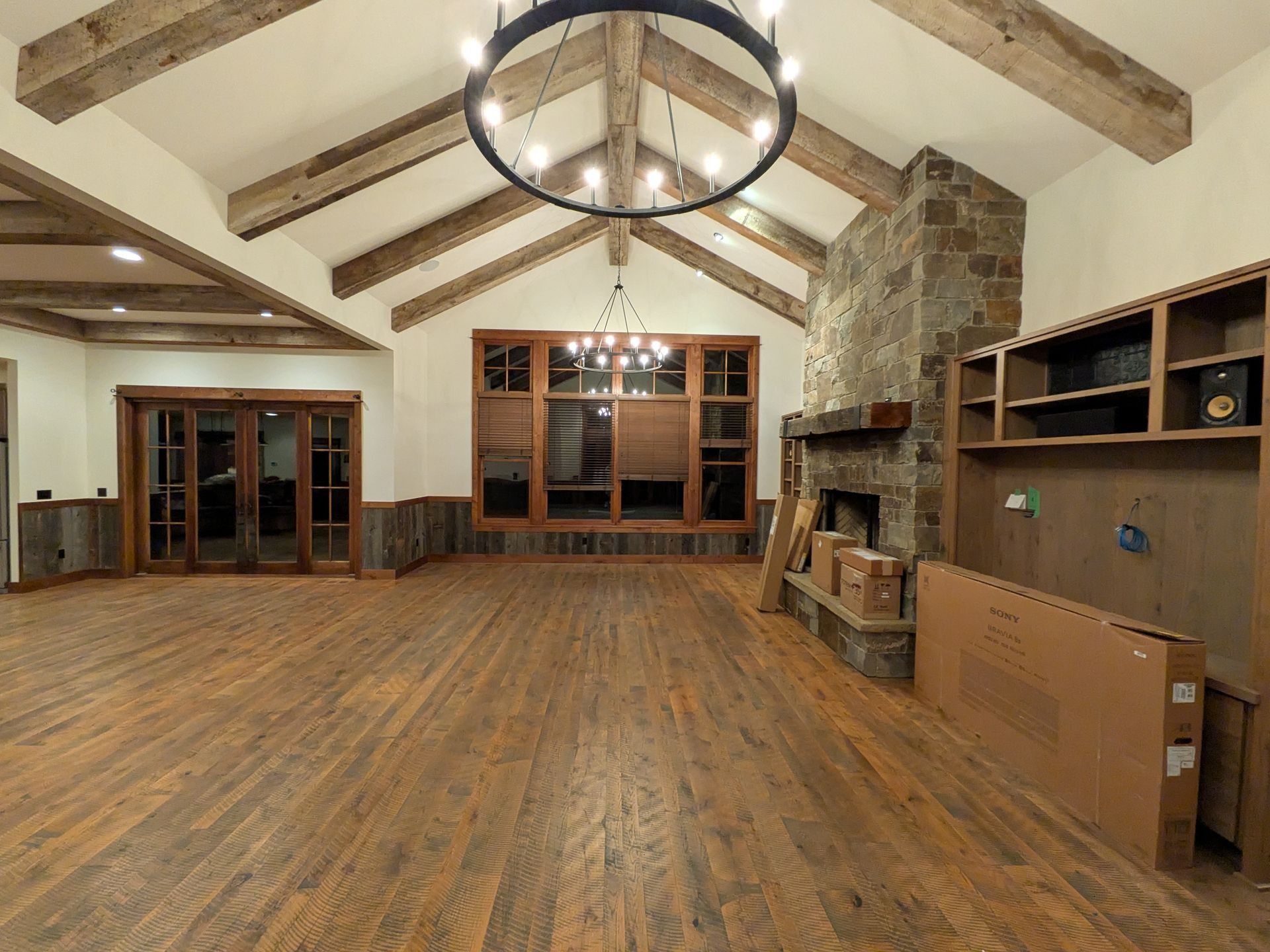 A spacious rustic living room with wood-beamed vaulted ceilings, herringbone floors, a stone fireplace, and a chandelier.