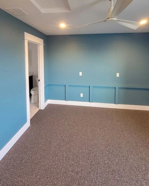 A room with blue walls, beige carpeting, white trim, a ceiling fan, and a doorway leading to a bathroom.