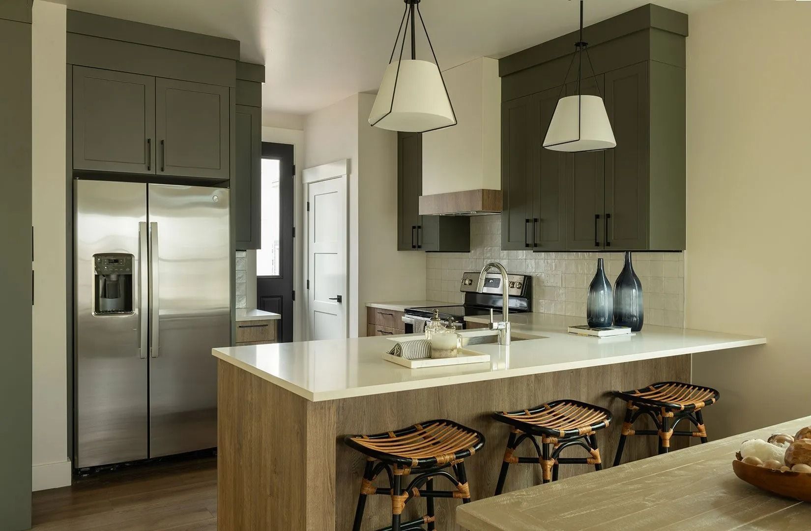 A modern kitchen with dark green cabinets, a wood-toned island with three stools, and a stainless steel refrigerator.