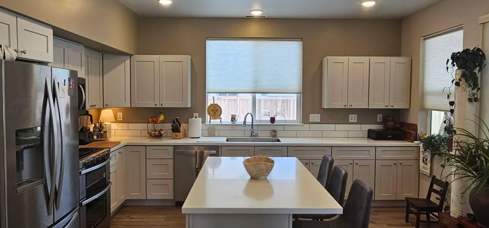 A kitchen with white cabinets, stainless steel appliances, a central island with bar seating, and windows with blinds.