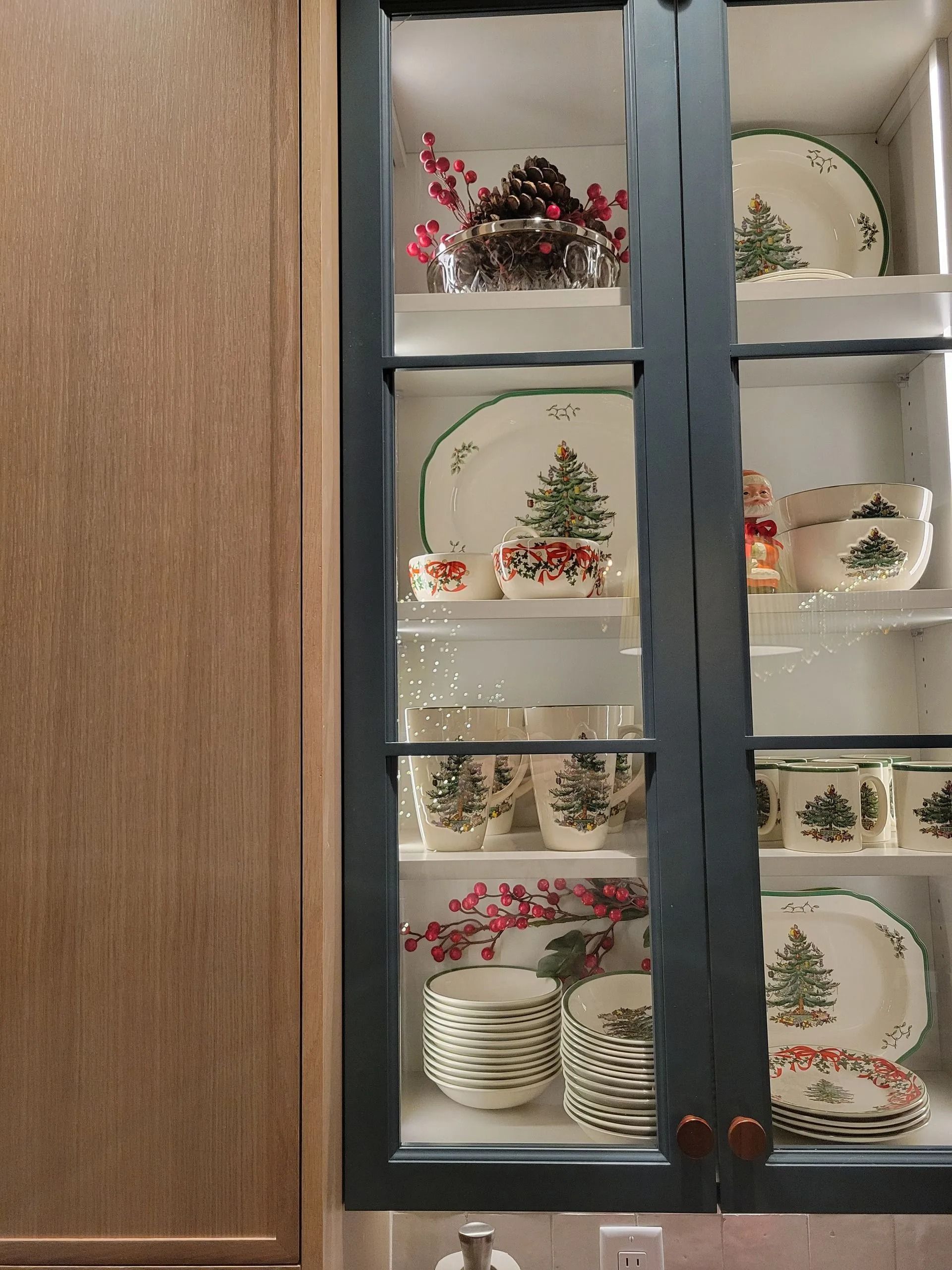 A glass-front cabinet with dark charcoal frames filled with Christmas-themed white dishes and festive holiday decor.