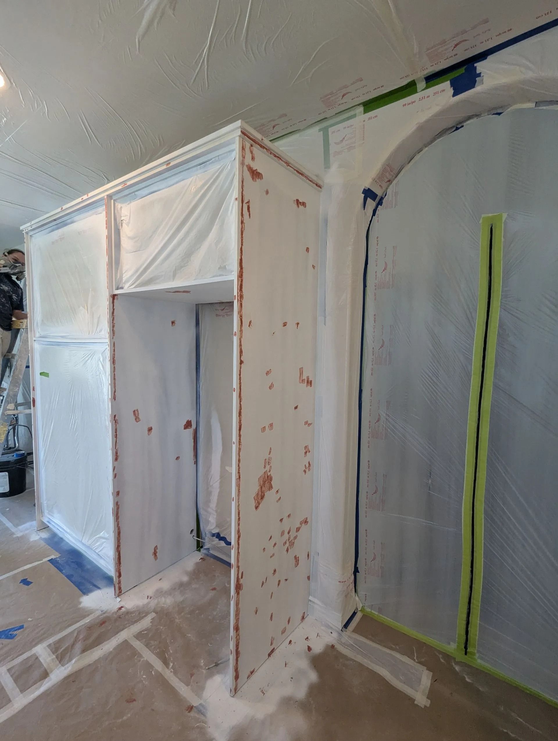 An interior space undergoing renovation, featuring cabinets covered in plastic film and an arched doorway masked off.